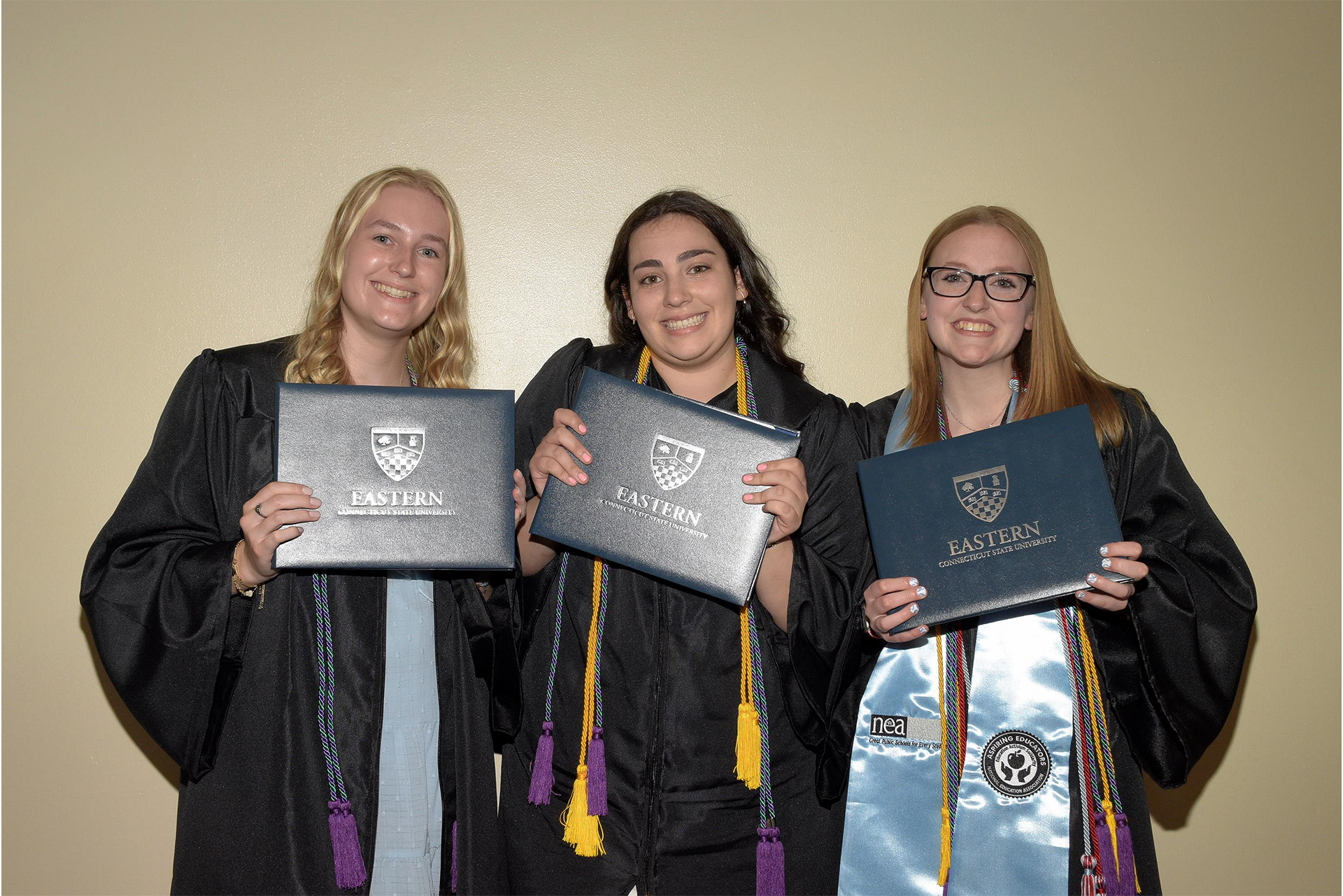 3 girls in graduation gowns holding diploma covers and smiling