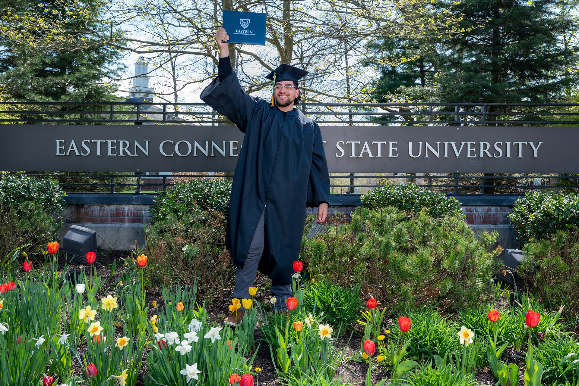 Student posing with diploma in front of Eastern's entrance sign