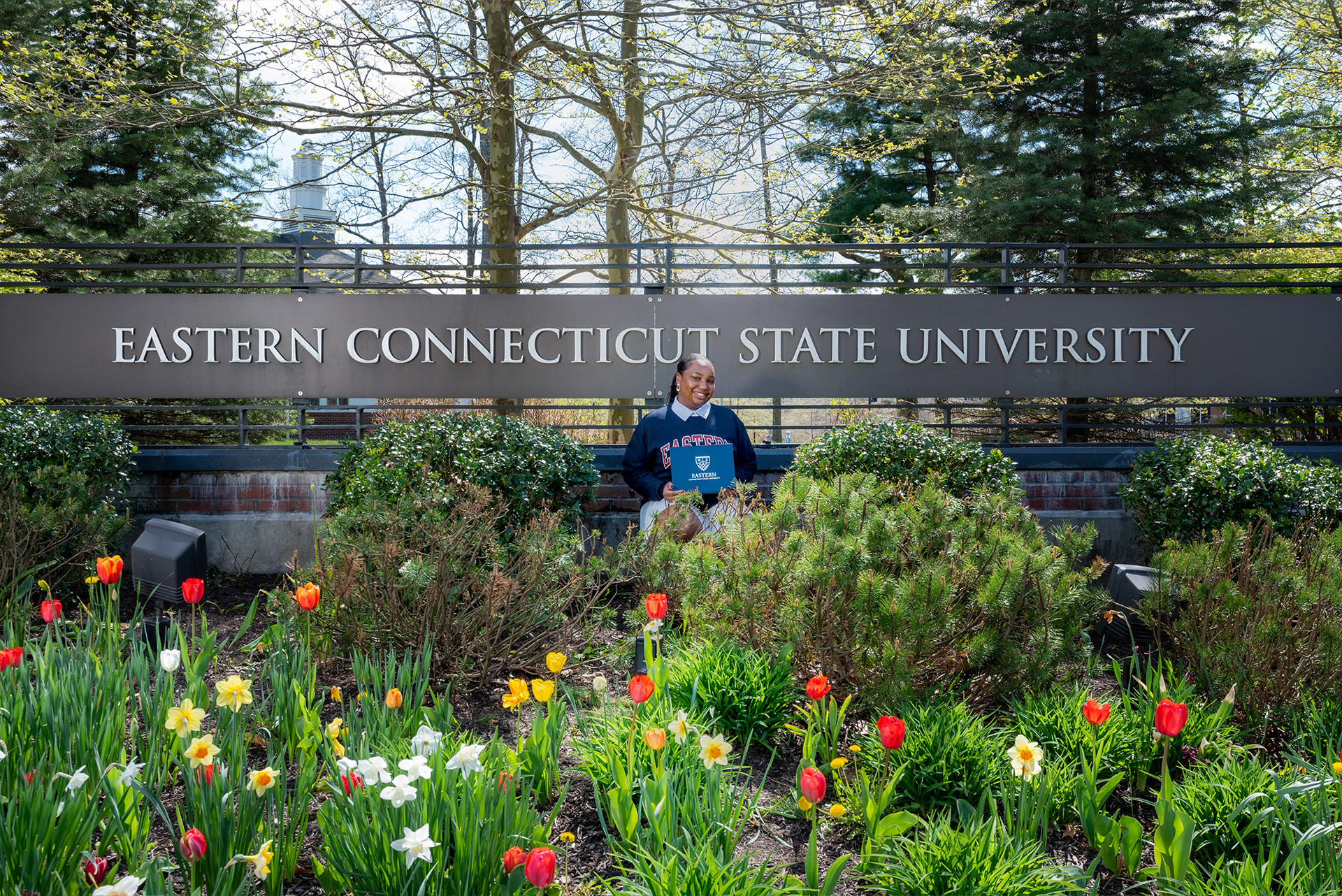 Student posing with diploma in front of Eastern's entrance sign